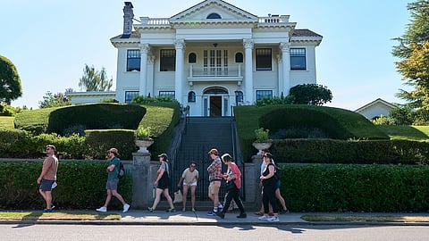 A group of seven people walks in front of a stately white house with columns as part of a theatrical tour called The Wealth Walk.