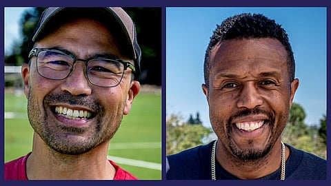 A man of Asian descent wears glasses, while a Black man wears a necklace and a T-shirt. Both men smile.