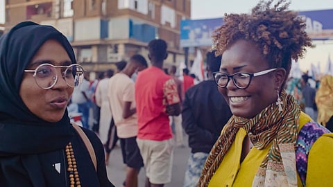 Two young Sudanese activists stand together smiling and speaking, not looking directly at the camera.