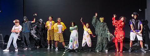Cast of the 2025 teen musical The Wiz poses on stage in vibrant, creative costumes representing various characters including animals, bricks, flowers, and fantastical figures, set against a dark theater backdrop.