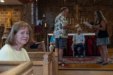 In the foreground, Margaret O'Donnell is seated in a pew and looking directly at the camera, while performers rehearse in the background at the front of the church, some sitting, some standing, all of them holding the script.