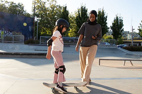Young girl in safety gear learning to skateboard at an outdoor skate park, guided by a woman in a hijab.