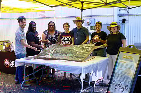 Group of seven people smiling under a yellow canopy at a Skyway Coalition community event, gathered around a 3D terrain model.