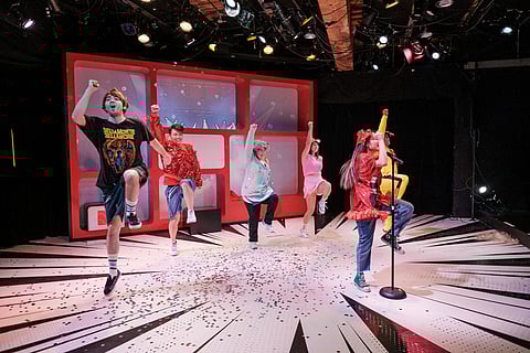 Actors perform an energetic dance routine on stage during a theatrical production, dressed in colorful costumes with confetti scattered across the floor under bright stage lighting.