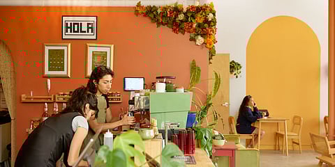 Baristas prepare drinks at Café Calaveras, a colorful Latino-owned coffee shop with orange and yellow walls, floral decorations, and plants, while a customer sits at a wooden table in the background.
