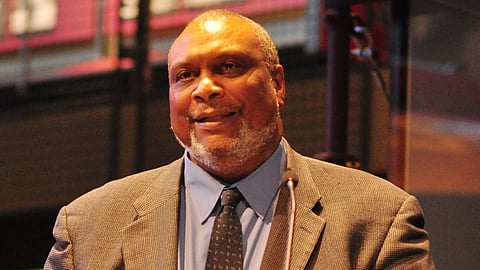 A Black man in a suit and tie speaking at an event, standing behind a microphone with a warm smile, captured in a close-up portrait under stage lighting.