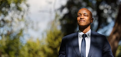 Portrait of Girmay Zahilay outdoors in a navy suit and tie, looking upward with a confident smile against a blurred background of trees and sky.