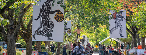 Large banners with Black art depicting figures carrying bundles hang between trees along a street. People walk, bike, and gather under the shade, with vendors and performers visible in the background.