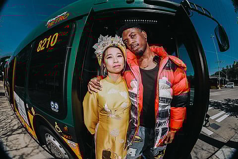 Two people stand arm in arm in front of a King County Metro bus. One wears a bright yellow áo dài with a decorative headdress, and the other wears a red and black puffer jacket with patterned pants, both looking directly at the camera.