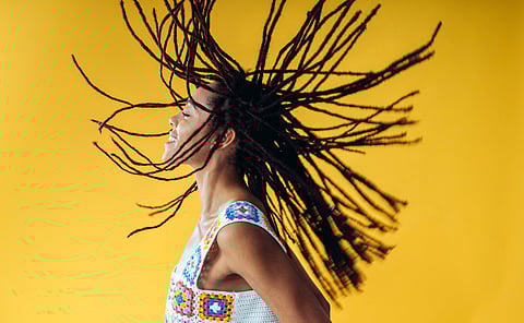 Smiling Black woman flipping long locs in motion, wearing a colorful crochet top against a vibrant yellow background.