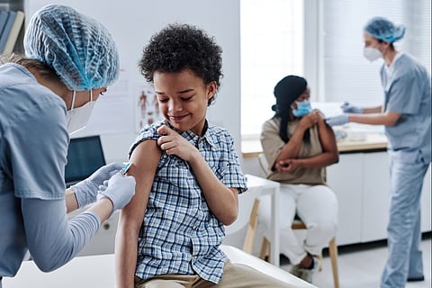 A Black child is vaccinated in the foreground while a Black adult is vaccinated in the background. The nurses are wearing masks, as is the Black adult.