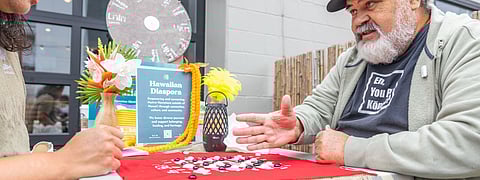 Two people play Kōnane at a decorated table featuring a Hawaiian Diaspora sign, flowers, and cultural elements; one player gestures mid-conversation.