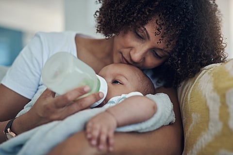 A Black mother gently feeds her infant from a bottle while cuddling on a couch.
