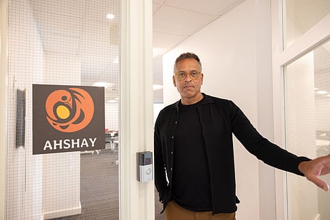 Middle-aged Black-presenting man standing at the entrance of an office with a sign reading “AHSHAY” featuring an orange and yellow abstract logo, wearing glasses and a black shirt, looking at the camera in a bright modern office setting.