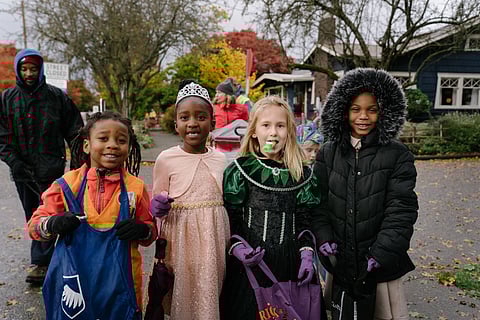Four children dressed in colorful costumes stand together outdoors, holding trick-or-treat bags and smiling. Autumn leaves cover the ground, and trees with fall foliage line the background of the neighborhood street.