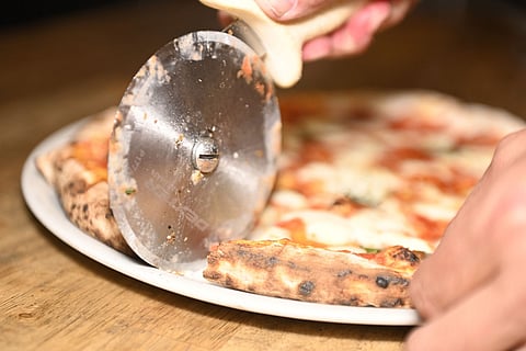 Close-up of a person cutting a freshly baked pizza with a metal pizza cutter on a white plate, showing melted cheese, tomato sauce, and a charred crust on a wooden table.
