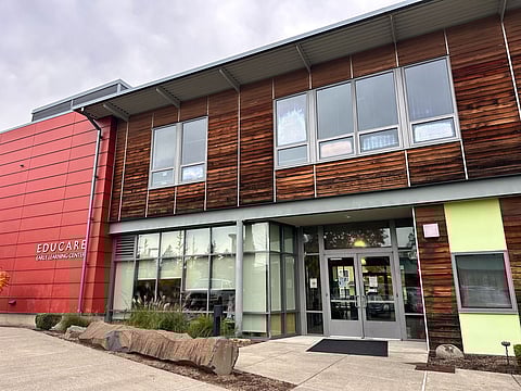 Modern building exterior of Educare Early Learning Center featuring wood paneling, red and green accent walls, large windows, and a glass double-door entrance under a covered awning on a cloudy day.