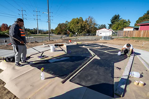 Two men work on painting a large outdoor mural shaped like a raised fist on the ground, using black paint and paper stencils at a community park site near a street and power lines on a sunny day.