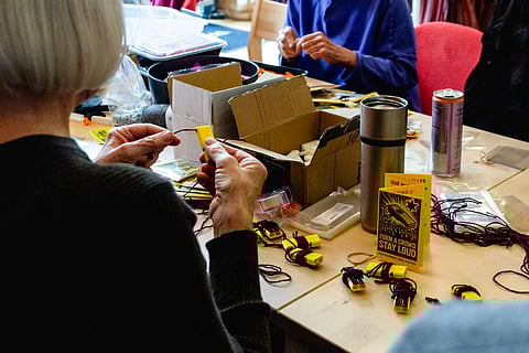 People assembling yellow protest whistles with purple cords around a table filled with craft materials, boxes, and a pamphlet reading 'Form a Crowd, Stay Loud.'