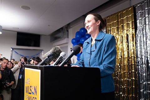 Smiling woman in a blue blazer speaks at a podium surrounded by microphones during a campaign event, with blue balloons, gold streamers, and a yellow ‘Katie Wilson for Mayor’ sign visible as supporters and photographers look on.