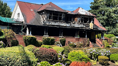 Burned two-story house with a charred roof and blackened walls, showing severe fire damage, surrounded by well-kept shrubs, hedges, and blooming hydrangeas under a bright blue sky.