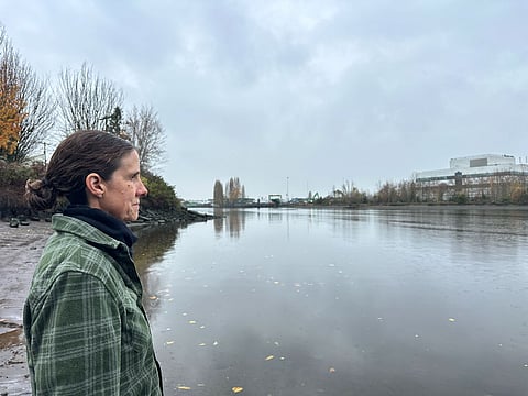 Person in a green plaid jacket standing on a muddy riverbank on an overcast day, looking across calm water toward distant trees and industrial buildings.