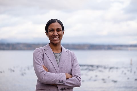A brown-skinned woman in a lilac suit jacket crosses her arms as she stands in front of a body of water.
