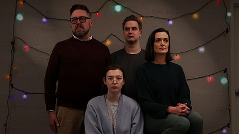 Family portrait–style image of four actors from the play “Life on the Moon,” posed against a wall with colorful string lights. Two stand in back and two sit in front, all with neutral expressions, representing a family in the production.