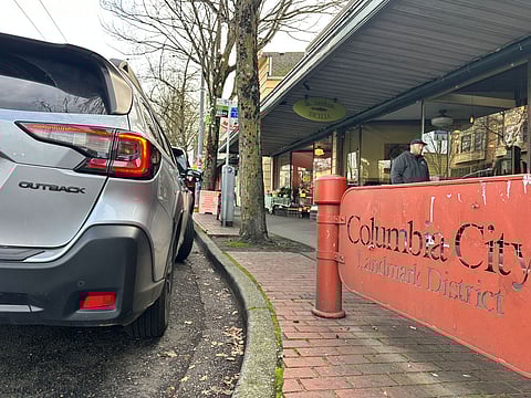 Columbia City Landmark District curbside with a parked silver Subaru Outback, a red district sign in the foreground, leafless trees, and storefronts along the sidewalk on a December day.