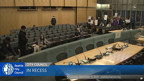 Protesters holding signs gather at the back of the Seattle City Council chambers as the meeting goes into recess, with a few attendees and officers scattered among mostly empty seats and vacant council desks.