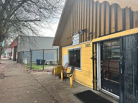 Smoke shop with yellow siding and barred windows on Rainier Avenue South, next to a fenced grassy lot and the gray L.E.M.S. building slated for redevelopment with Jazz Night School.