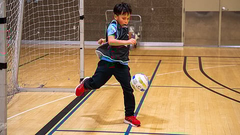 James Parrish, 7, learns futsal skills; he is standing on a basketball court in front of a goal and is mid-kick of the ball.