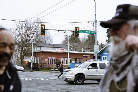 Activists Eddie Rye and Bob Barnes stand at the corner of Martin Luther King Jr. Way South and South Cherry Street. Behind them is an intersection with a sign that reads Martin Luther King Jr. Vehicles and people move across the intersection.