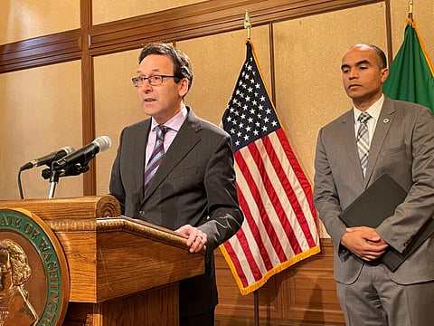 Washington Gov. Bob Ferguson speaks at a podium during a press conference, with Attorney General Nick Brown standing beside him, U.S. flag behind them, discussing state preparations for potential federal immigration enforcement actions.