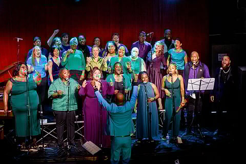Social-justice choir Resounding Love sings on stage during the “Love Will Win” concert at Black & Tan Hall, led by a conductor, with diverse members performing into microphones under warm stage lighting.