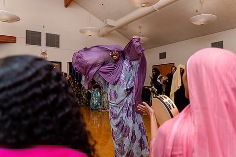A smiling, young Somali woman wearing glasses and a purple headscarf performs a traditional Somali dance surrounded by a circle of girls and young women. One woman in the circle plays a tambourine.