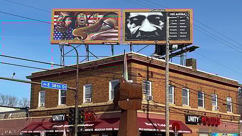 George Floyd Square at the intersection of East 38th Street and Chicago Avenue in South Minneapolis. A statue of a Black Power fist is in the foreground and billboards above the building show George Floyd and an illustration of Black people grieving.