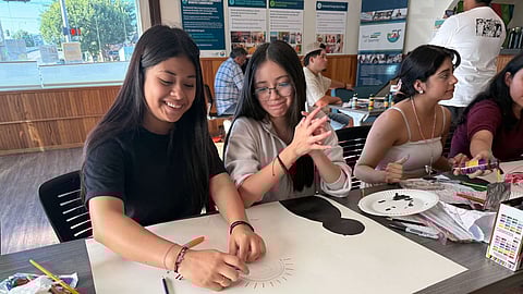 Two youth create art on a large piece of paper set on a table with art supplies all around them. Other youth are to the side and in the background, also making art.