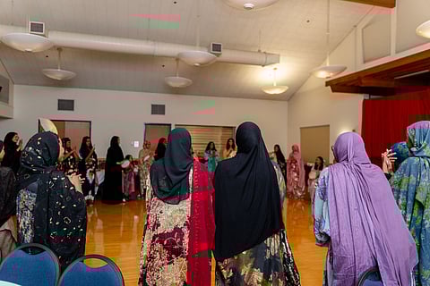 Women wearing colorful hijabs and traditional dresses gathered in a community hall, standing in a circle and clapping.