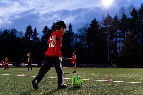 Young soccer player in red jersey practicing with a green soccer ball on an outdoor turf field at dusk, with teammates and field lights in the background.