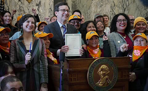Washington state official holds a signed document at a podium with the Seal of Washington, surrounded by supporters wearing yellow hats and orange bandanas.