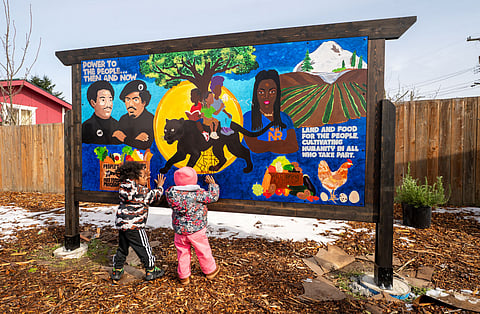 Two children in winter jackets stand in front of a colorful community mural inspired by the Black Panther Party, featuring portraits, a black panther, farmland, and messages about “Power to the People” and food justice.