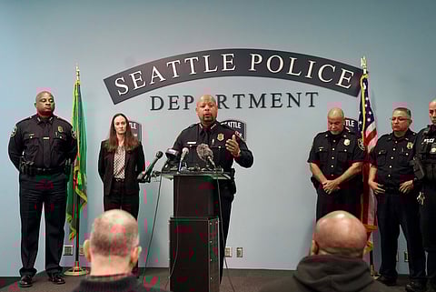 Seattle Police Department press conference with a uniformed police chief speaking at a podium with microphones, flanked by other officers and Mayor Katie Wilson, with U.S. and Washington state flags behind them and “Seattle Police Department” signage on the wall.