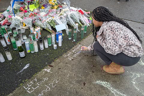 A Black teen crouches writing a chalk message beside a street memorial with candles, flowers, and notes, honoring a victim at a community vigil.