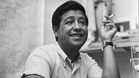 Cesar Chavez smiles while holding a cigarette at a desk.