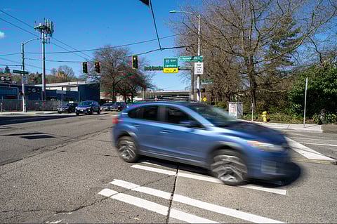 Vehicles move along Rainier Avenue South near South Massachusetts Street, south of the I-90 overpass. The traffic light in the shot is red, and the car in the forefront is moving to the right of the frame and is blurry.