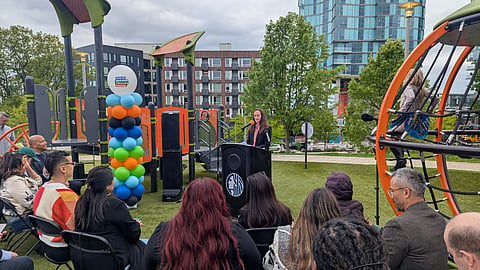 Woman speaking at a podium during an outdoor press event at a modern playground.