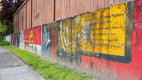 Colorful memorial mural painted on a concrete wall beside a sidewalk, featuring a dedication to fallen community members.