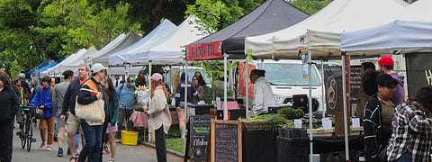 Crowds browse vendor tents at Columbia City Farmers Market in Seattle under grey skies.