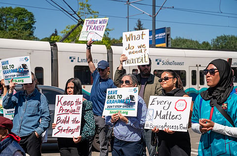 Poeple at a rally hold up signs in support of transit access and light rail; a light rail train is in the background.